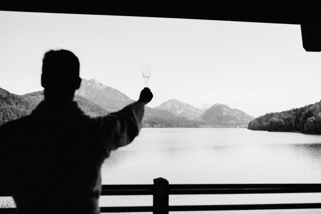 Ein Hochzeitsgast hebt sein Glas vor dem Panorama des Fuschlsees